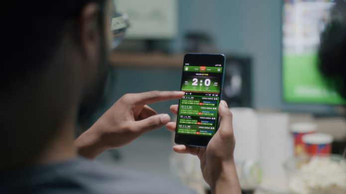 Una persona que mira un partit de futbol sosté un mòbil on es pot veure una app d'apostes esportives