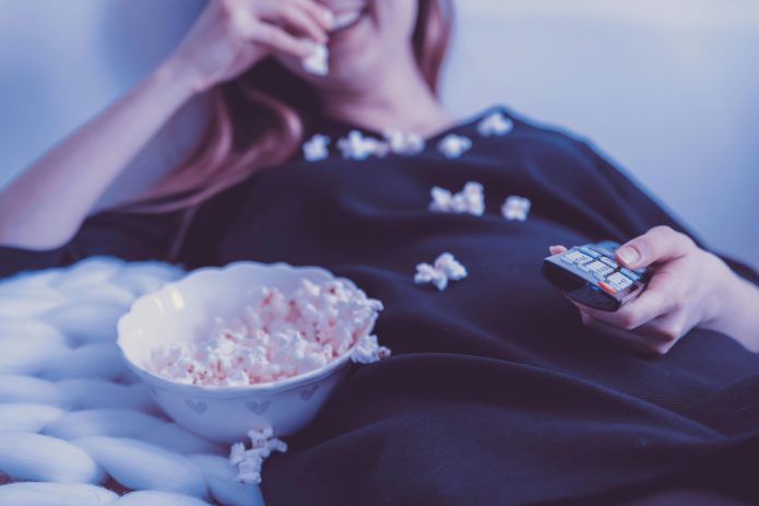 Persona mirant una sèrie en streaming amb crispetes i comandament a distància, representant el binge watching i el consum intensiu de plataformes digitals.